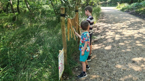 Two young boys playing wind chimes with sticks, a path and trees in the background.